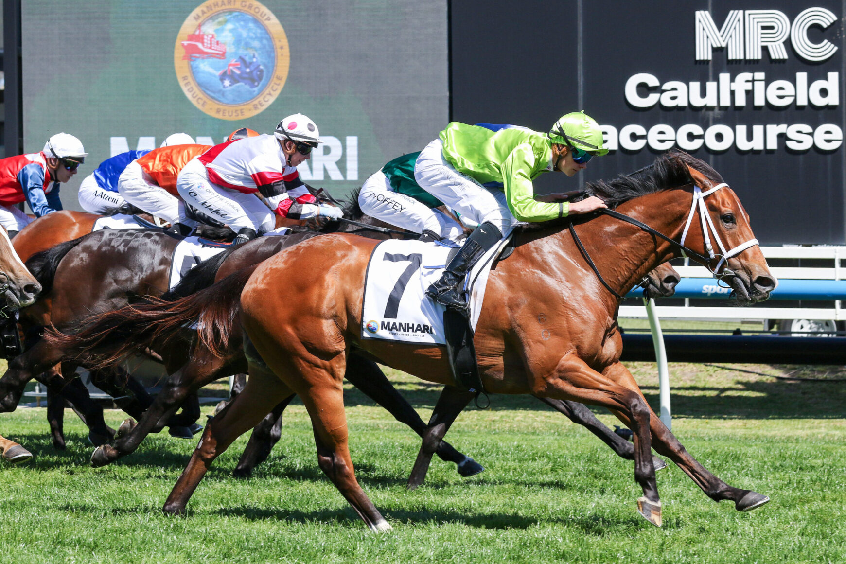 Thoroughbred race in progress; leading chestnut horse with lime-green silks rider, others close behind on the grass track.