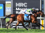 Two racehorses with jockeys in colorful silks race side by side in front of a large MRC sign at the track café.