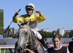 Jockey in yellow-and-white silks celebrates on a gray horse, raising a fist as a handler holds the reins beside them at a racecourse.