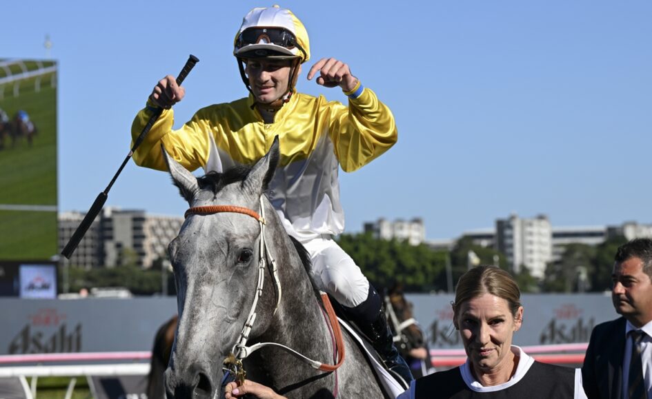 Jockey in yellow-and-white silks celebrates on a gray horse, raising a fist as a handler holds the reins beside them at a racecourse.