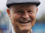 Close-up of a smiling older man wearing a gray flat cap, outdoors, showing warm facial wrinkles.