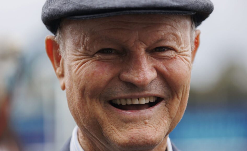 Close-up of a smiling older man wearing a gray flat cap, outdoors, showing warm facial wrinkles.