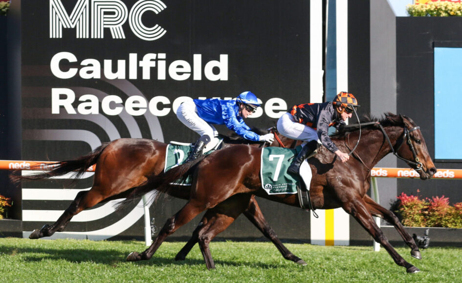 Two jockeys on dark brown horses sprint toward the finish at Caulfield Racecourse, race banners in the background.