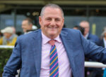 Smiling man in a blue checkered suit with a pink shirt and striped tie outdoors, arms spread for a friendly pose.