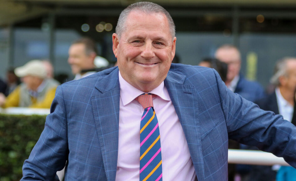 Smiling man in a blue checkered suit with a pink shirt and striped tie outdoors, arms spread for a friendly pose.