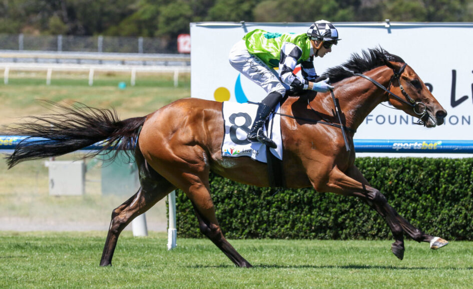 Jockey wearing a neon green vest rides a brown racehorse mid-race on a grassy track.