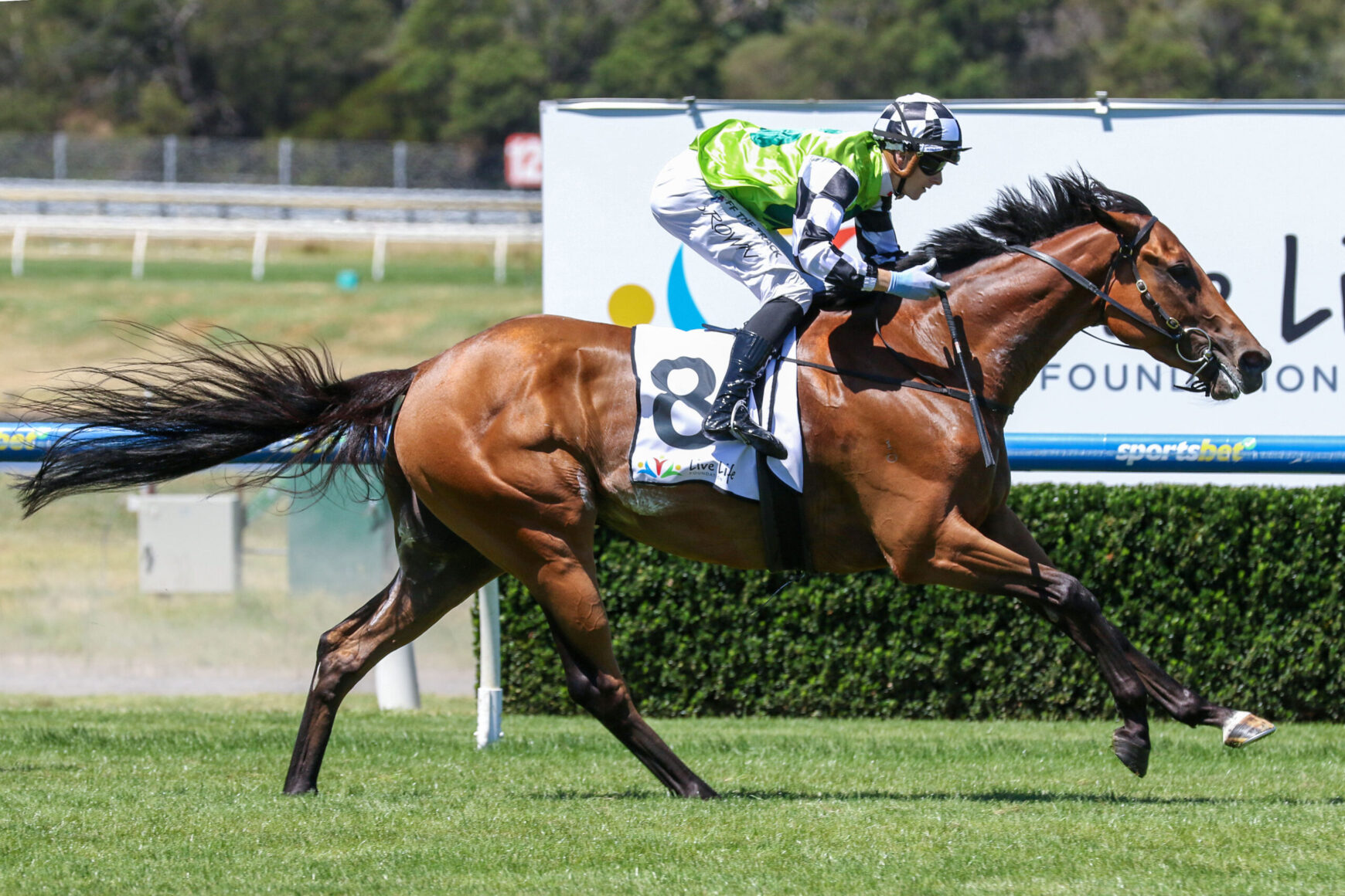 Jockey wearing a neon green vest rides a brown racehorse mid-race on a grassy track.