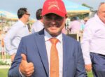 Man in a blue suit gives a thumbs-up at an outdoor event, wearing a red Magic Millions cap.