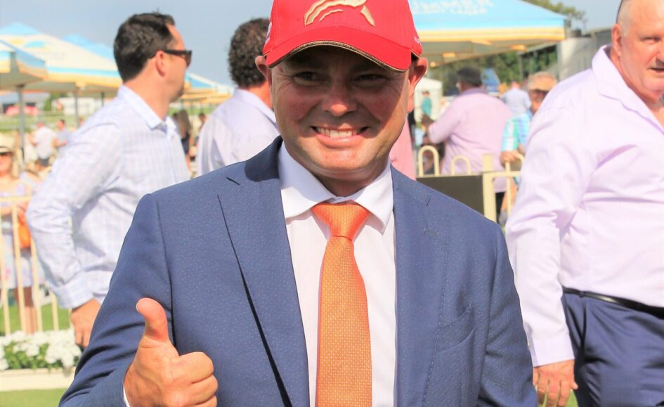 Man in a blue suit gives a thumbs-up at an outdoor event, wearing a red Magic Millions cap.