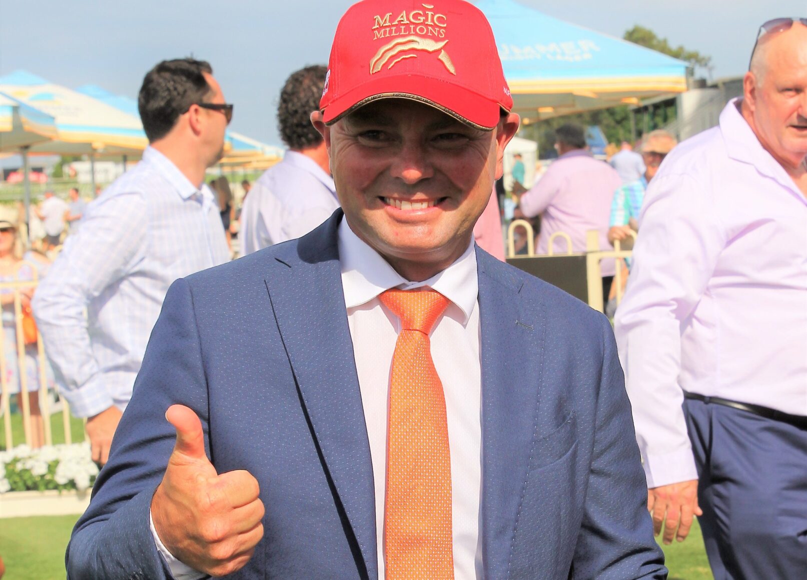 Man in a blue suit gives a thumbs-up at an outdoor event, wearing a red Magic Millions cap.