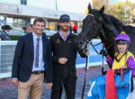 Three men pose with a racehorse and jockey at a racetrack; one in a suit, one in a Sparta Racing jacket, the jockey in purple-yellow silks stands nearby.