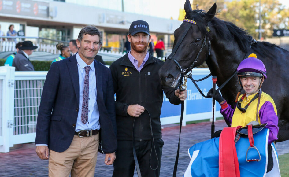 Three men pose with a racehorse and jockey at a racetrack; one in a suit, one in a Sparta Racing jacket, the jockey in purple-yellow silks stands nearby.
