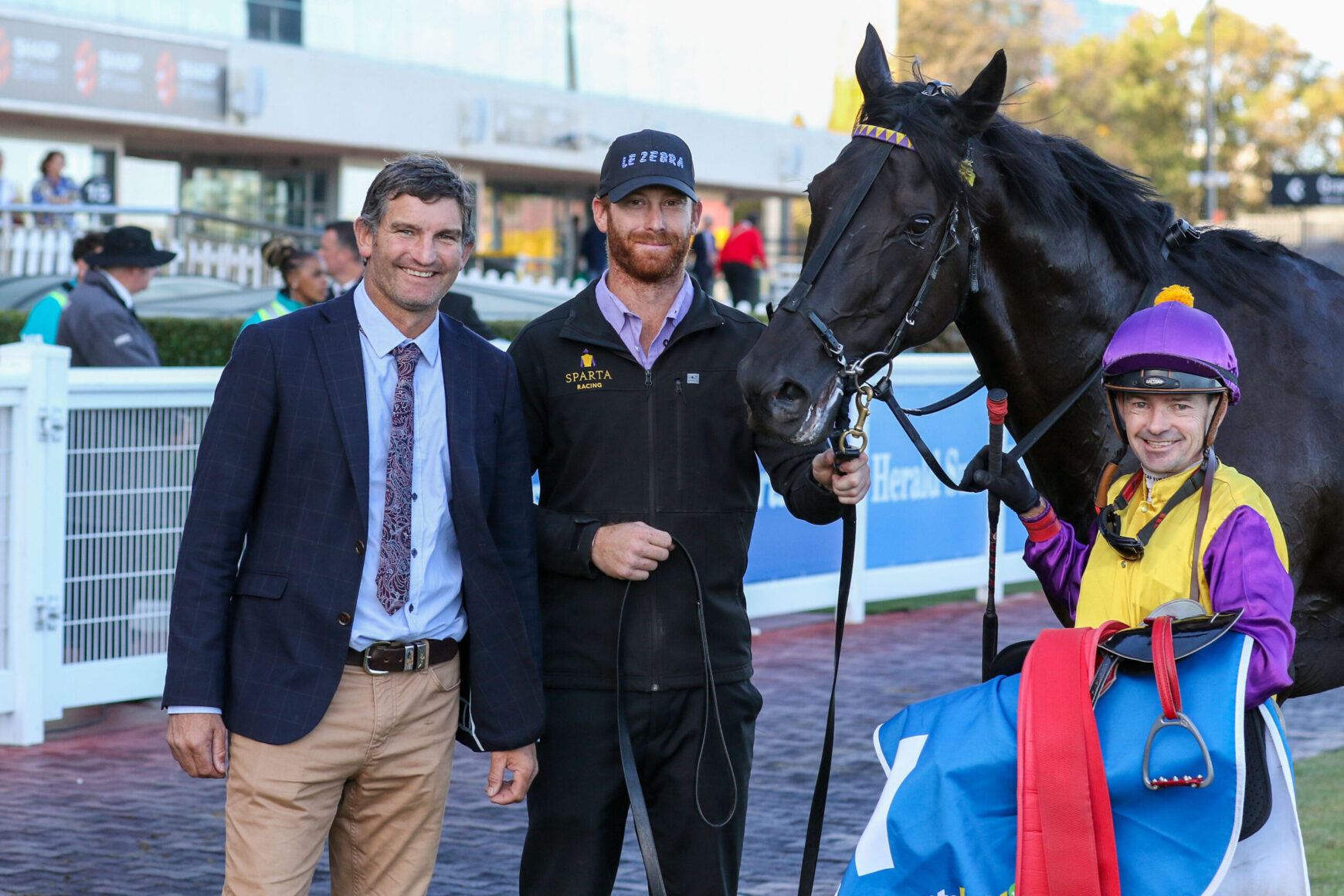 Three men pose with a racehorse and jockey at a racetrack; one in a suit, one in a Sparta Racing jacket, the jockey in purple-yellow silks stands nearby.