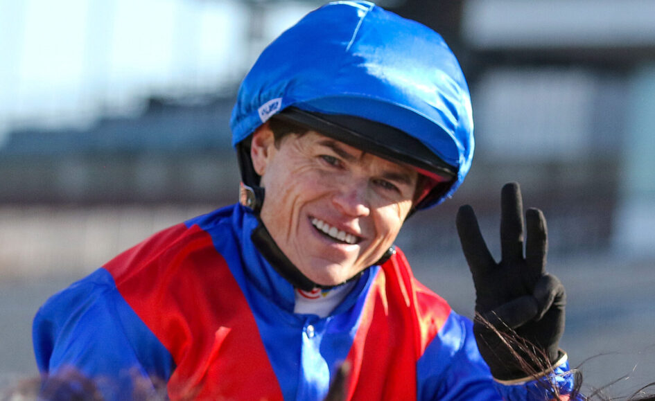 Smiling jockey in blue helmet and red-and-blue silks, flashing a peace sign with gloved hand.