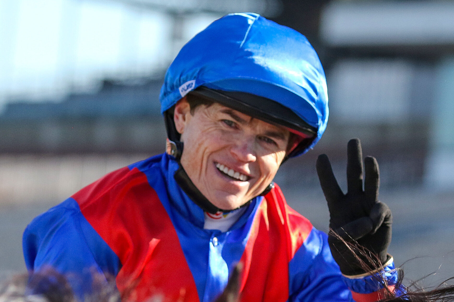 Smiling jockey in blue helmet and red-and-blue silks, flashing a peace sign with gloved hand.