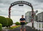 Man standing with arms crossed under a flower-covered arch that reads 'Eagle Farm' with cloudy sky and modern buildings in the background.