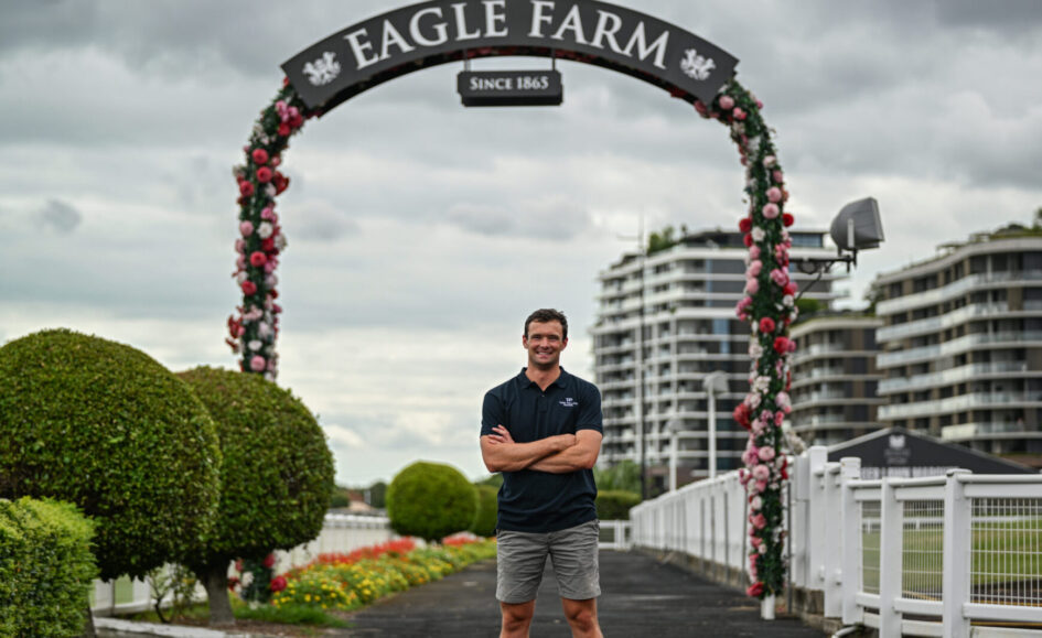 Man standing with arms crossed under a flower-covered arch that reads 'Eagle Farm' with cloudy sky and modern buildings in the background.