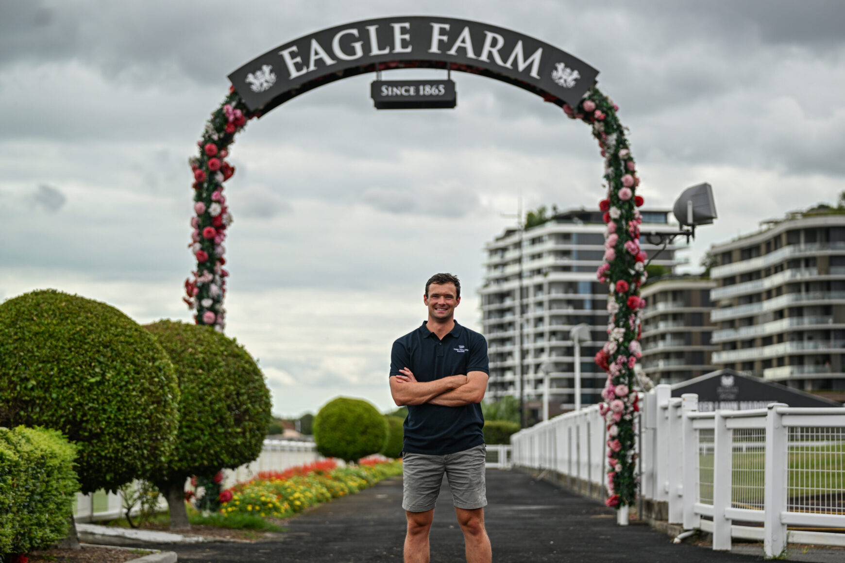 Man standing with arms crossed under a flower-covered arch that reads 'Eagle Farm' with cloudy sky and modern buildings in the background.