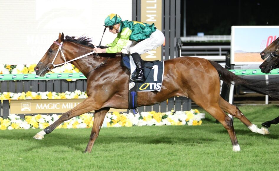 Jockey in green/yellow silks riding a brown horse with saddlecloth number 1 during a race, finish-line setup and yellow flowers in the background.