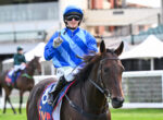 Jockey in bright blue striped silks and helmet riding a brown horse at a race track, pointing upward with gloved hand.