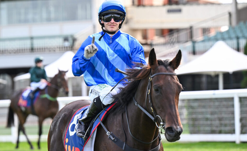 Jockey in bright blue striped silks and helmet riding a brown horse at a race track, pointing upward with gloved hand.