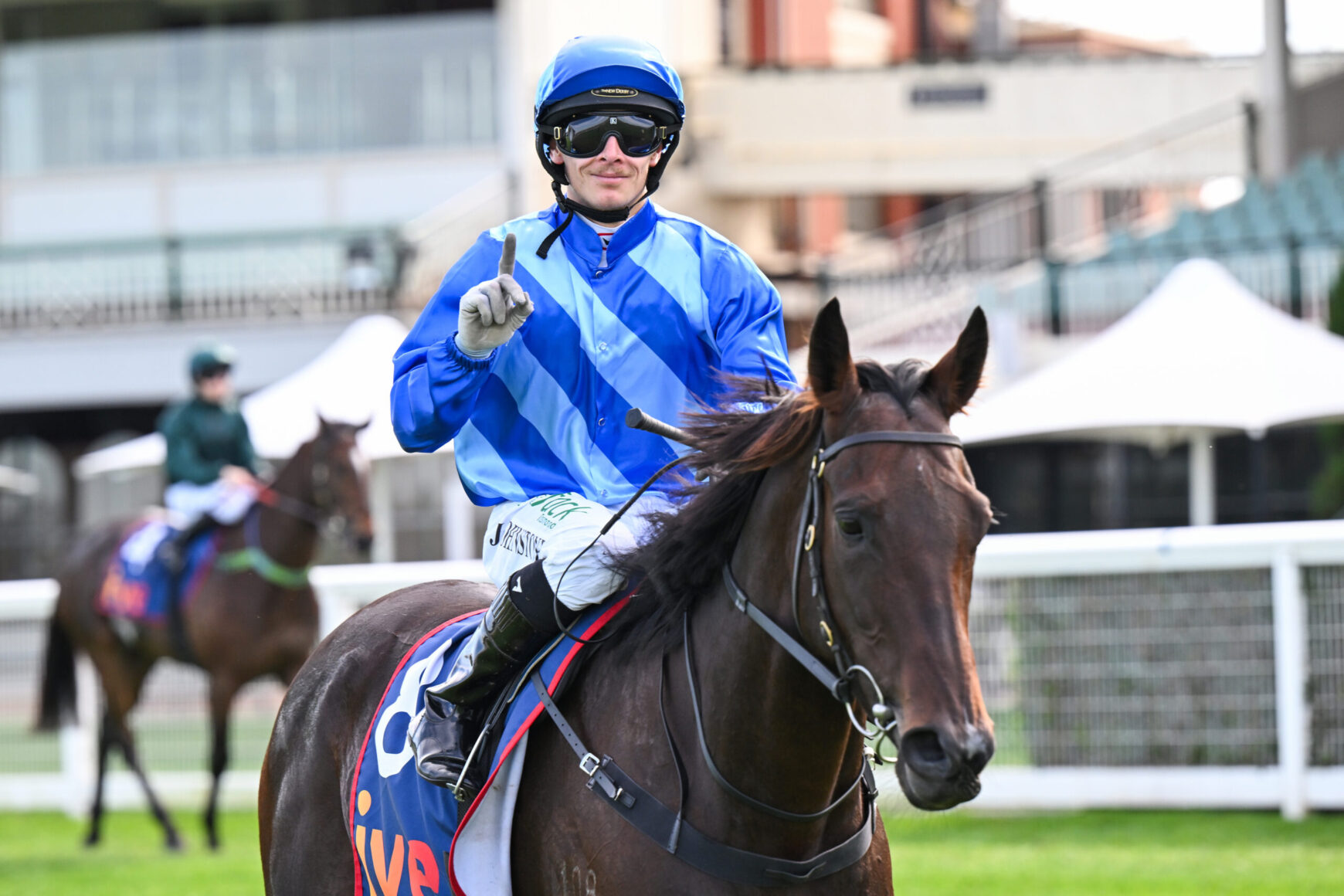 Jockey in bright blue striped silks and helmet riding a brown horse at a race track, pointing upward with gloved hand.