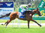 Bay racehorse with rider in green-and-white silks speeding along a grassy track past billboards and rails on a sunny day.