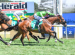 Jockeys on racing horses sprint toward the finish line, green and white silks, number 7 visible on the lead horse, on a grassy track.