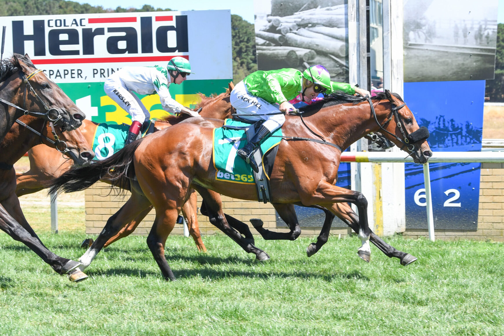 Jockeys on racing horses sprint toward the finish line, green and white silks, number 7 visible on the lead horse, on a grassy track.