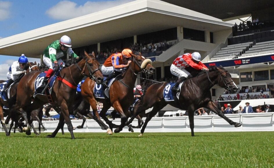 Group of jockeys in colorful silks racing on a green turf track with a modern grandstand and spectators in the background