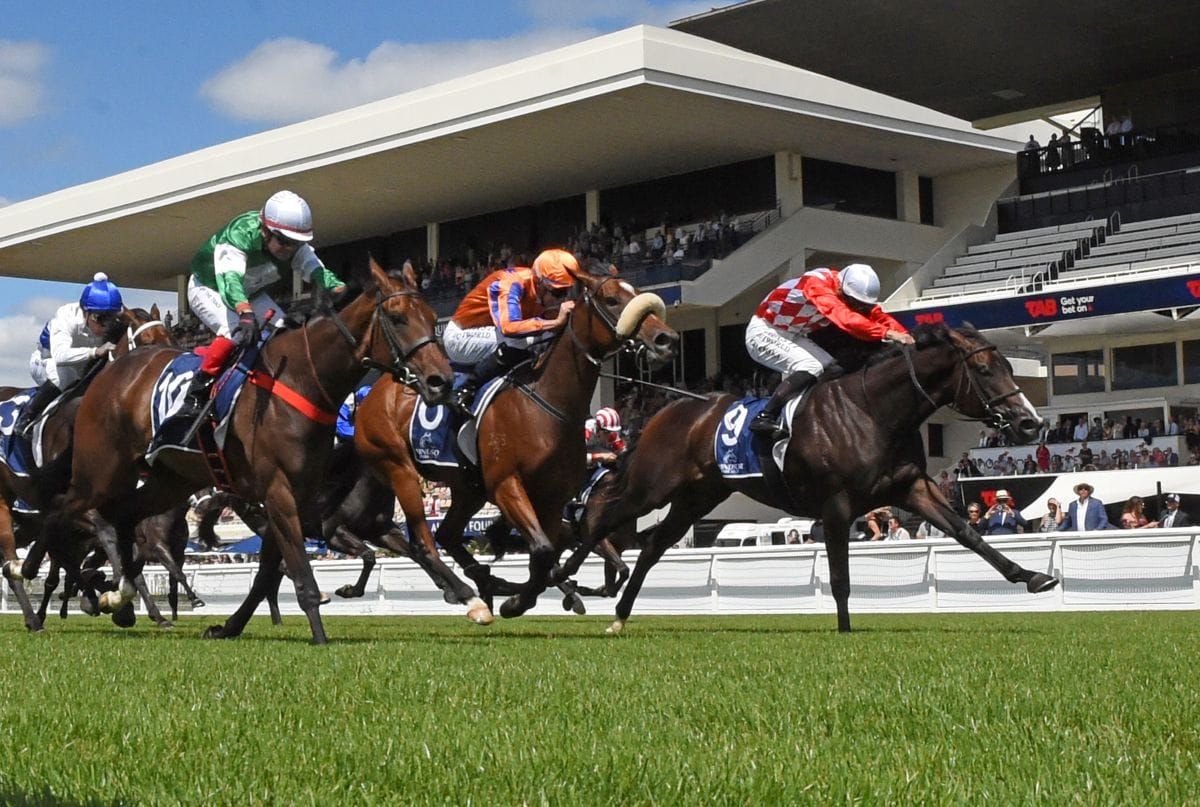 Group of jockeys in colorful silks racing on a green turf track with a modern grandstand and spectators in the background