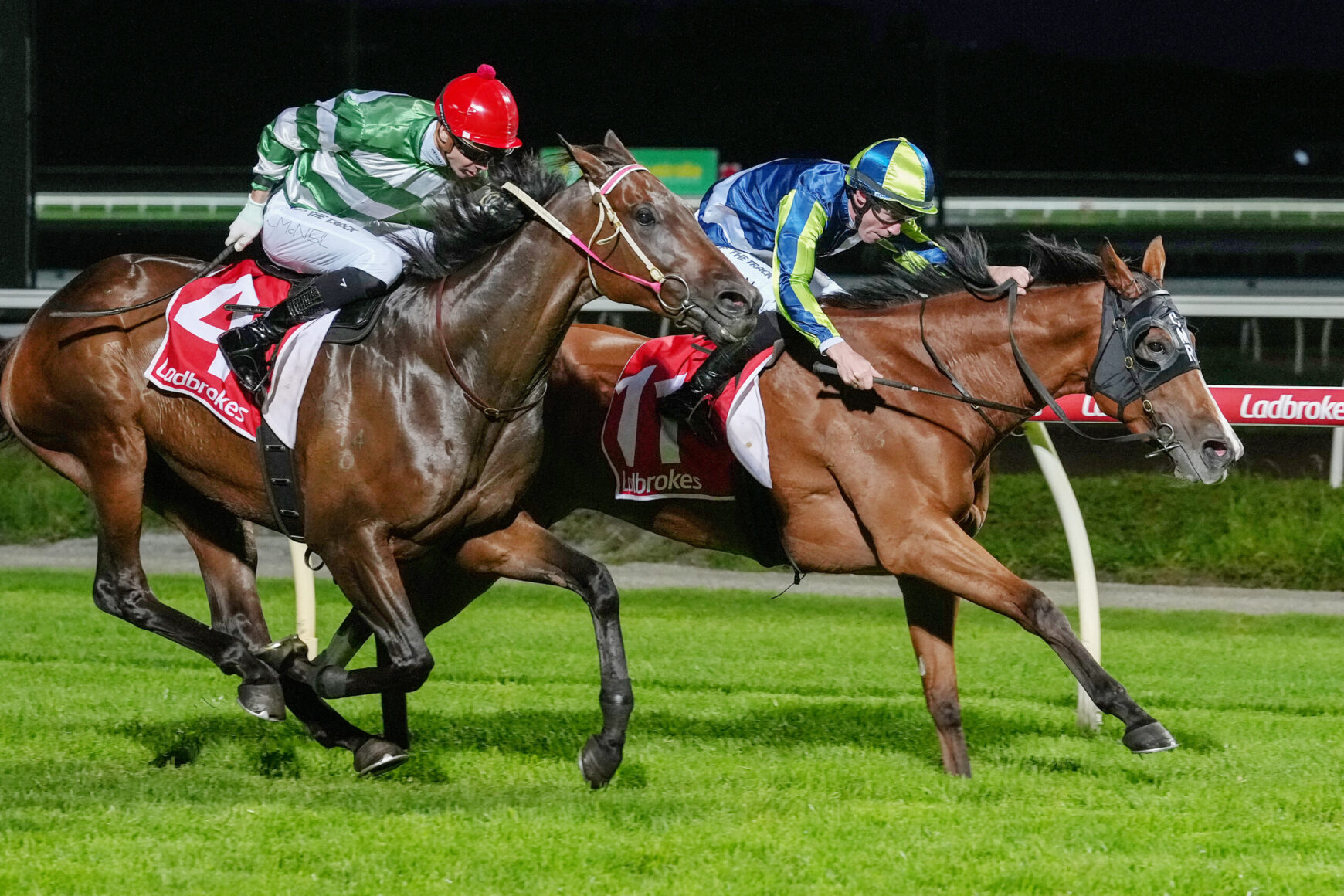 Two jockeys ride closely in a nighttime horse race, leaning forward as these sprinting horses stride on the turf with a Ladbrokes banner visible on the saddleship.
