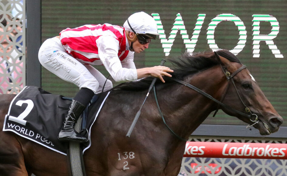 Jockey in white helmet and red-and-white silks leans forward on a dark brown horse during a race, saddle cloth shows number 2 with Ladbrokes signage in the background to the right.