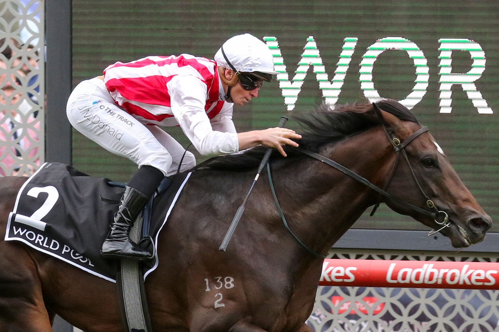 Jockey in white helmet and red-and-white silks leans forward on a dark brown horse during a race, saddle cloth shows number 2 with Ladbrokes signage in the background to the right.