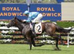Jockey in light blue silks rides a dark brown horse with number 2 on a racecourse, mid-stride at a sprint.