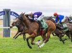 Jockeys race side by side on sprinting horses at a grassy track, with the lead horse wearing purple and white silks.