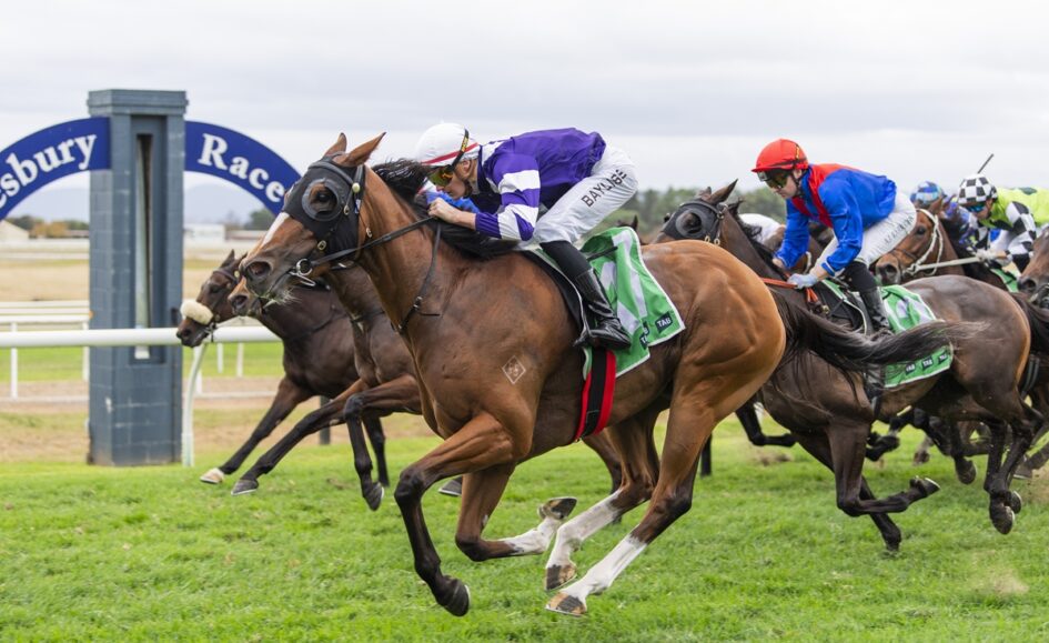 Jockeys race side by side on sprinting horses at a grassy track, with the lead horse wearing purple and white silks.