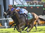Three jockeys on galloping horses neck-and-neck during a race at a sunny racetrack with a scoreboard behind them.