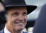 Close-up of a smiling man wearing a black hat and suit outdoors, looking to the right