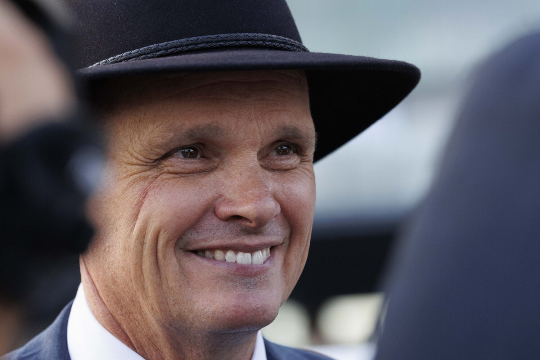 Close-up of a smiling man wearing a black hat and suit outdoors, looking to the right