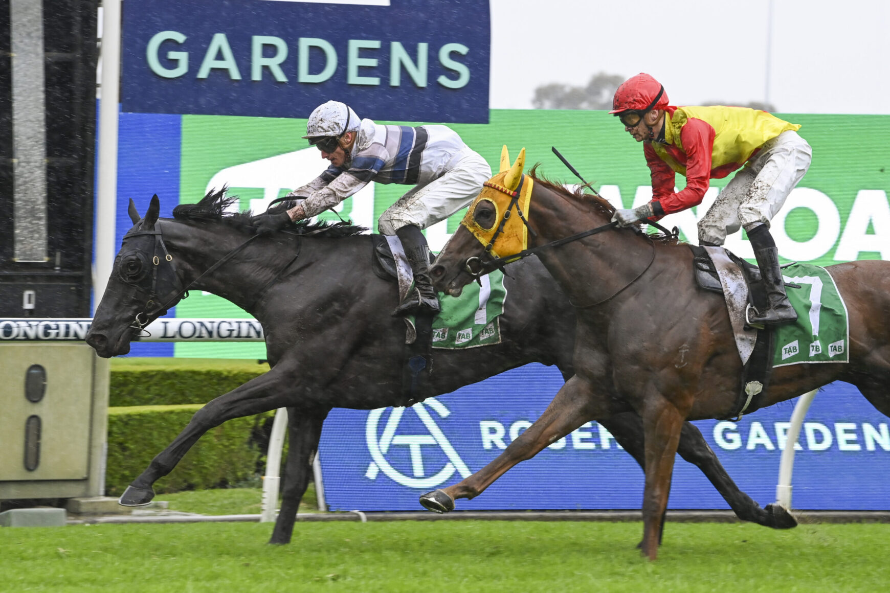 Two jockeys race on muddy horses, leaning forward as they sprint past green grass with a large GARDENS sign in the background.