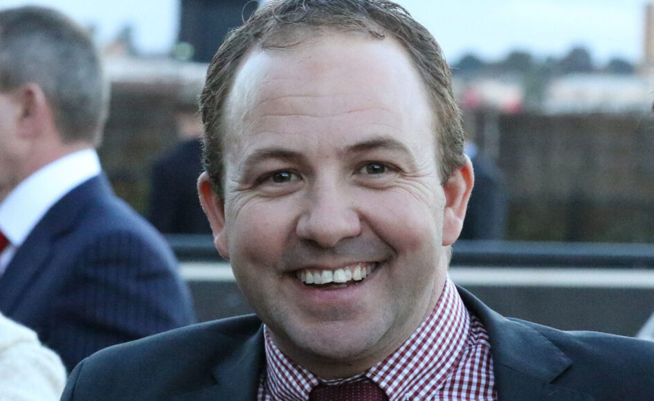 Smiling man in a navy suit with a red checkered shirt at an outdoor event, close-up portrait.