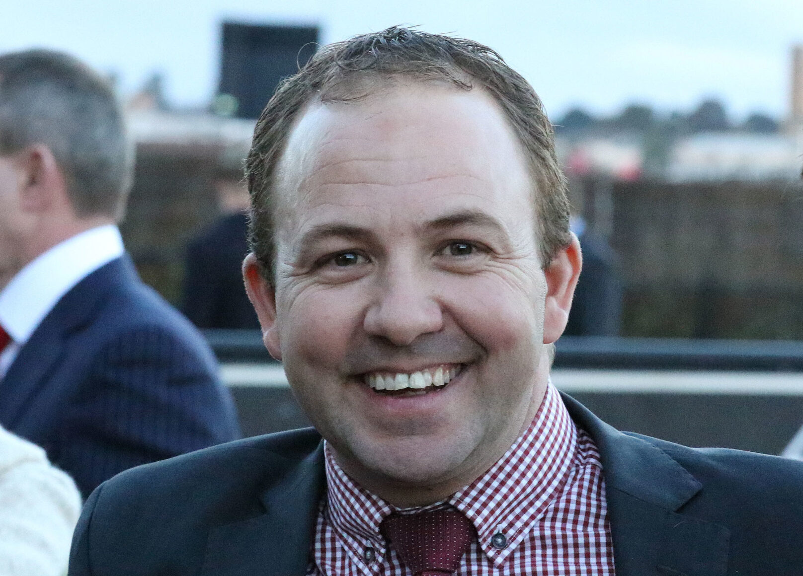 Smiling man in a navy suit with a red checkered shirt at an outdoor event, close-up portrait.