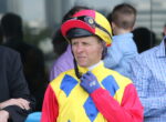 Jockey in yellow, blue, and red racing silks with a pink-yellow helmet, adjusting his blue glove and looking to the left.