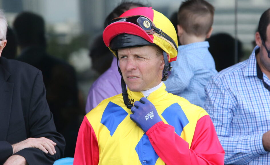 Jockey in yellow, blue, and red racing silks with a pink-yellow helmet, adjusting his blue glove and looking to the left.