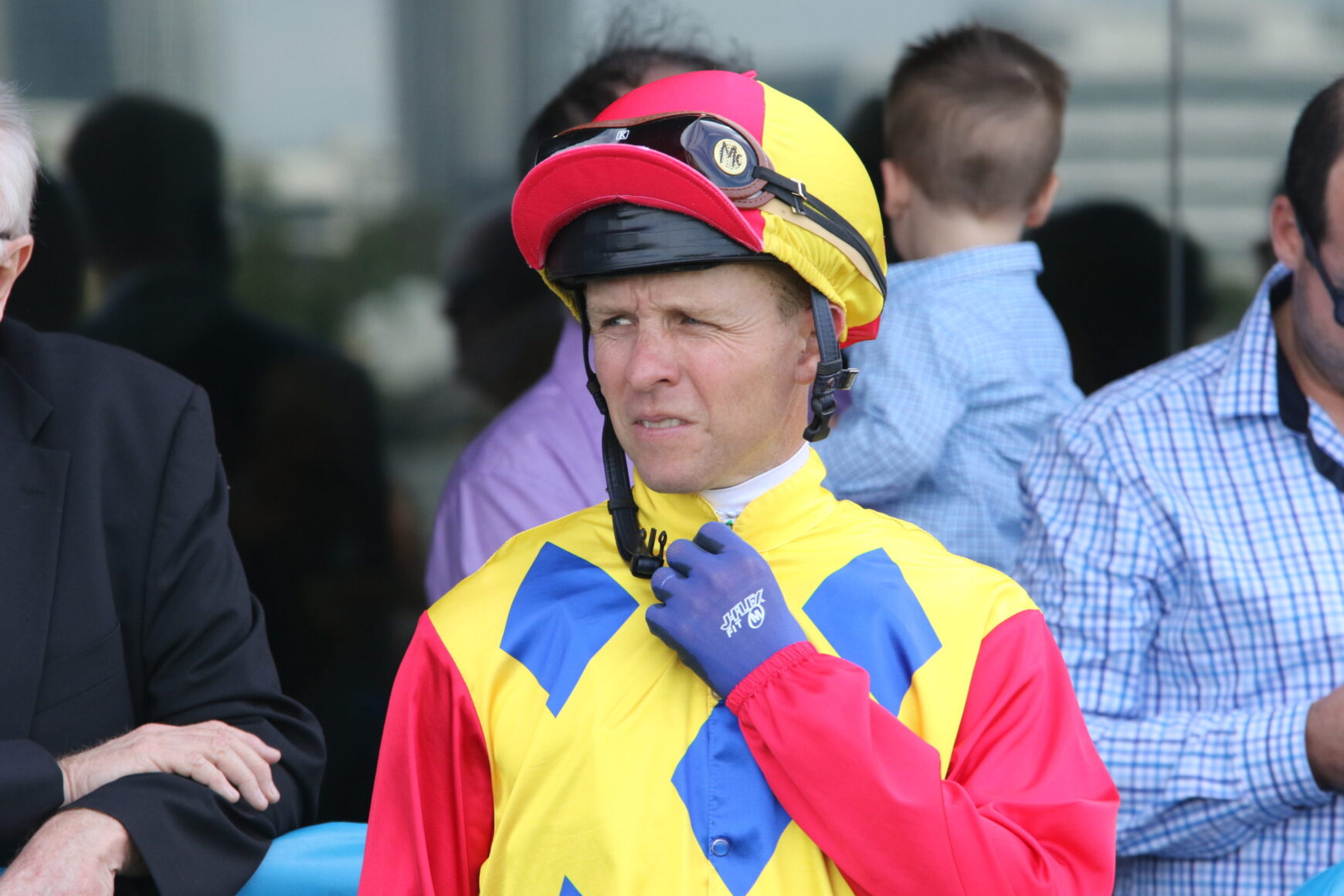 Jockey in yellow, blue, and red racing silks with a pink-yellow helmet, adjusting his blue glove and looking to the left.