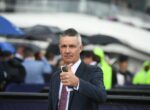 Man in a dark suit giving a thumbs-up and holding a smartphone, at a rainy outdoor event with blurred spectators and umbrellas in the background