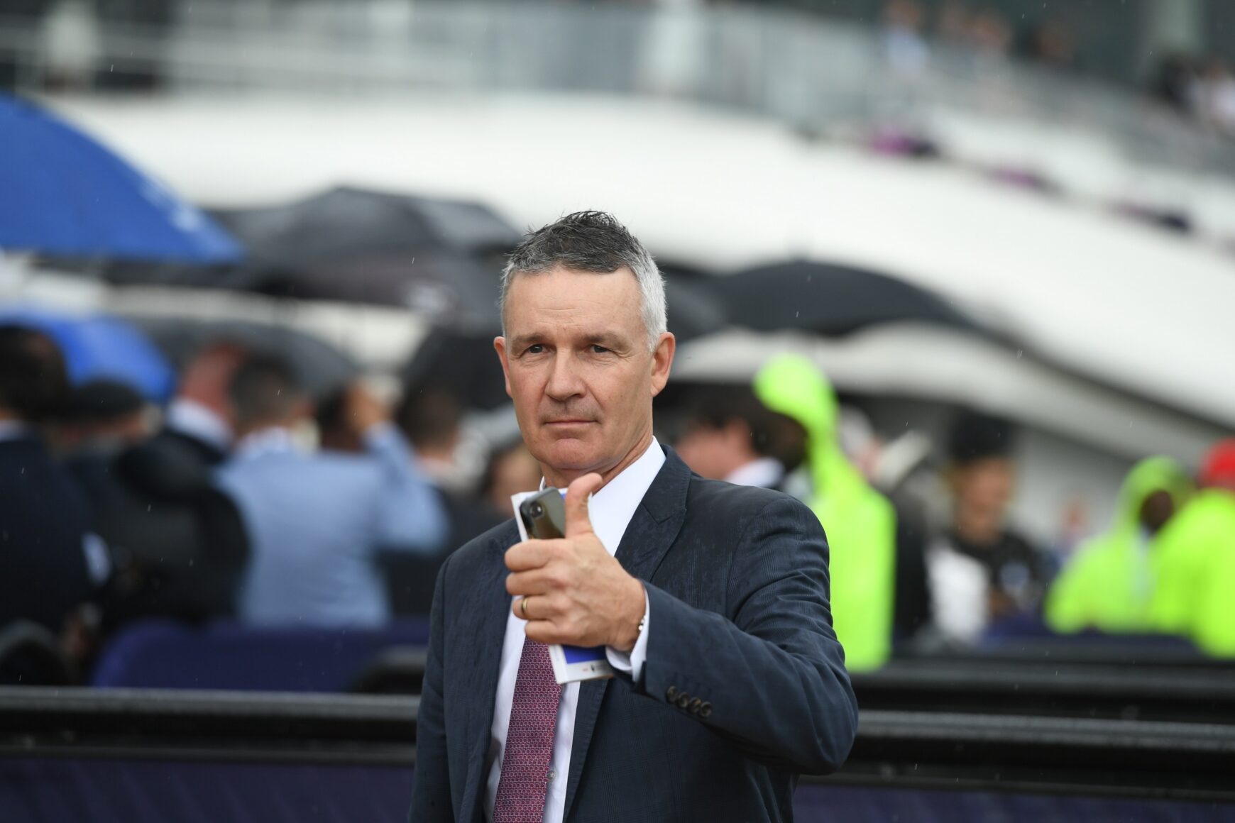Man in a dark suit giving a thumbs-up and holding a smartphone, at a rainy outdoor event with blurred spectators and umbrellas in the background