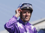 Smiling jockey in purple racing silks adjusts his helmet with STAR on the visor outdoors at a track.