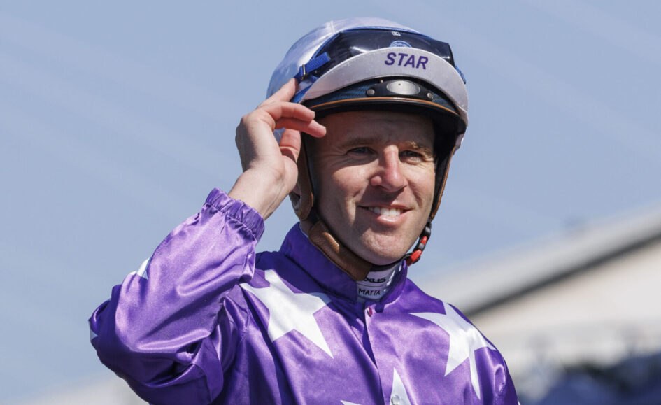 Smiling jockey in purple racing silks adjusts his helmet with STAR on the visor outdoors at a track.